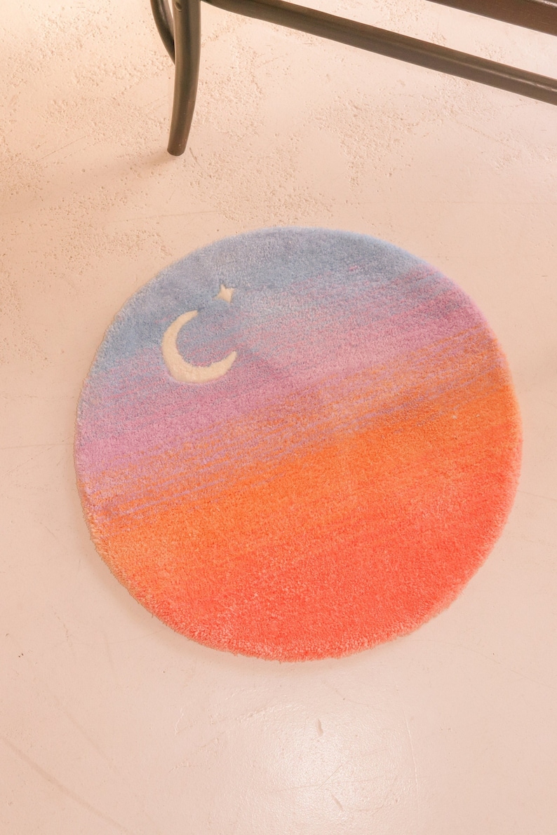 May include: Round rug with a gradient design in shades of blue, pink, and orange, resembling a sunset. A white crescent moon and star are featured in the blue section. The rug is a soft, plush material.