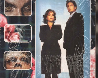 May include: A VHS tape cover for the X-Files episode "X-posed". The cover features a photo of the two main characters, Mulder and Scully, standing side-by-side. The title "The X-Files" is written in large, bold letters at the top of the cover. The episode title "X-posed" is written in smaller letters below the main title. The cover also includes the text "Written by Michael Joseph Edited by David Richter".