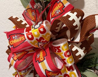 May include: A decorative wreath featuring a large multi-layered bow made of red, white, and yellow ribbon printed with the Kansas City Chiefs logo and text, interspersed with brown football-patterned ribbon, accented by green eucalyptus leaves.