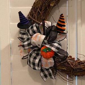 Könnte beinhalten: Eine schwarz-weiße karierte Schleife mit dem Text "TRICK OR TREAT" und "HAUNTED" darauf. Die Schleife ist mit einem kleinen orangefarbenen Kürbis und zwei schwarzen und orangefarbenen Hexenhüten verziert. Die Schleife ist an einem Weinrebenkranz befestigt.