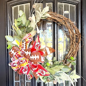 Puede incluir: Una corona decorativa con una base de ramas de vid naturales de color marrón, adornada con hojas de eucalipto verdes y un gran lazo de varias capas. El lazo presenta logotipos de los Kansas City Chiefs y gráficos de balones.