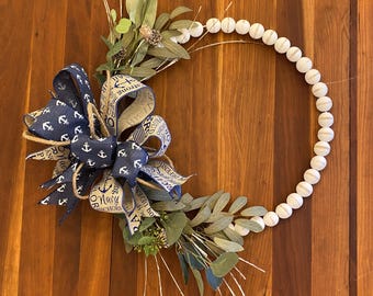 May include: A white beaded wreath with a blue and white ribbon bow with the text "Navy Anchors" and "Strong & Proud" on it. The wreath is decorated with green leaves.