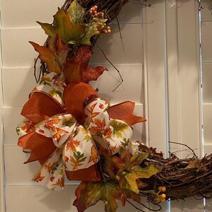 May include: A seasonal wreath with a grapevine base, decorated with orange and white ribbons printed with autumn leaves. The wreath also features artificial fall leaves in orange, green, and red, and small orange berries.