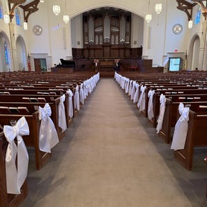 Puede incluir: El interior de un santuario de iglesia con filas de bancos de madera oscura dispuestos a ambos lados de un pasillo central. Cada extremo del banco está decorado con un gran lazo de cinta de satén blanco. Un gran órgano está al frente.