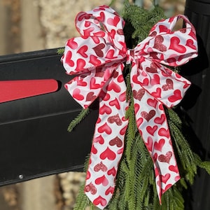 May include: A red and white bow with a heart pattern, tied to a green evergreen branch. The bow is made of satin ribbon and has a shiny finish.