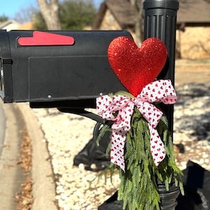 May include: A black mailbox decorated with a red glitter heart, a white bow with red hearts, and green sprigs.