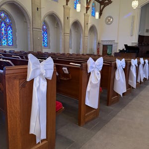 Könnte beinhalten: Eine Reihe von hölzernen Kirchenbänken, die mit großen, weißen Satinschleifen an den Enden verziert sind. Die Schleifen haben lange, fließende Stoffbänder, die an der Seite jeder Bank herabhängen. Der Innenraum zeigt Buntglasfenster in Blau.