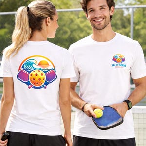 May include: White t-shirts with a colorful pickleball design. The shirts feature a logo with a wave, sun, and pickleball paddles. One shirt has the text "Salty Serve Academy". A person is holding a pickleball and paddle.