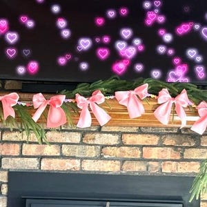 May include: A pink satin ribbon garland with bows hangs above a brick fireplace. The garland is decorated with small, colourful beads. The fireplace is decorated with a TV screen displaying a background of pink hearts.
