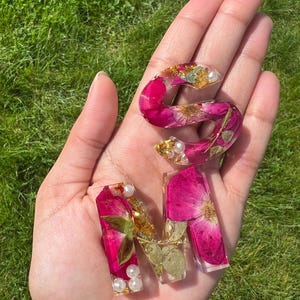 May include: Clear resin letters spelling the word "MOM". The letters are filled with pink flower petals, gold flakes, and small white pearls. The letters are displayed against a background of green grass.