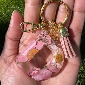 May include: A clear resin letter "D" keychain filled with pink and yellow dried flowers. The keychain has a gold-colored clasp, keyring, and tassel. The tassel is a light pink color.