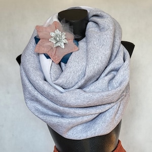 May include: A gray infinity scarf with a decorative flower pin. The scarf is made of a soft, textured fabric and is draped around a mannequin head. The flower pin has a pink and silver design with blue accents.