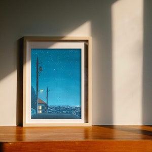May include: Framed art print depicting a nighttime cityscape under a starry sky. The artwork features a street with a lamppost, a house with lit windows, and a distant city skyline. The print is in a light wooden frame.
