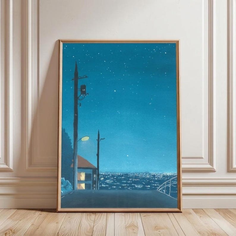 May include: A framed art print depicts a nighttime cityscape. The artwork features a road, a house with lit windows, and utility poles under a starry, teal-colored sky. The print is in a wooden frame and leans against a white wall.