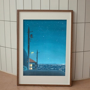 May include: Framed art print depicting a nighttime cityscape under a starry sky. The artwork features a road leading towards a town with illuminated buildings. A house with lit windows and utility poles are in the foreground. The frame is a warm brown color.