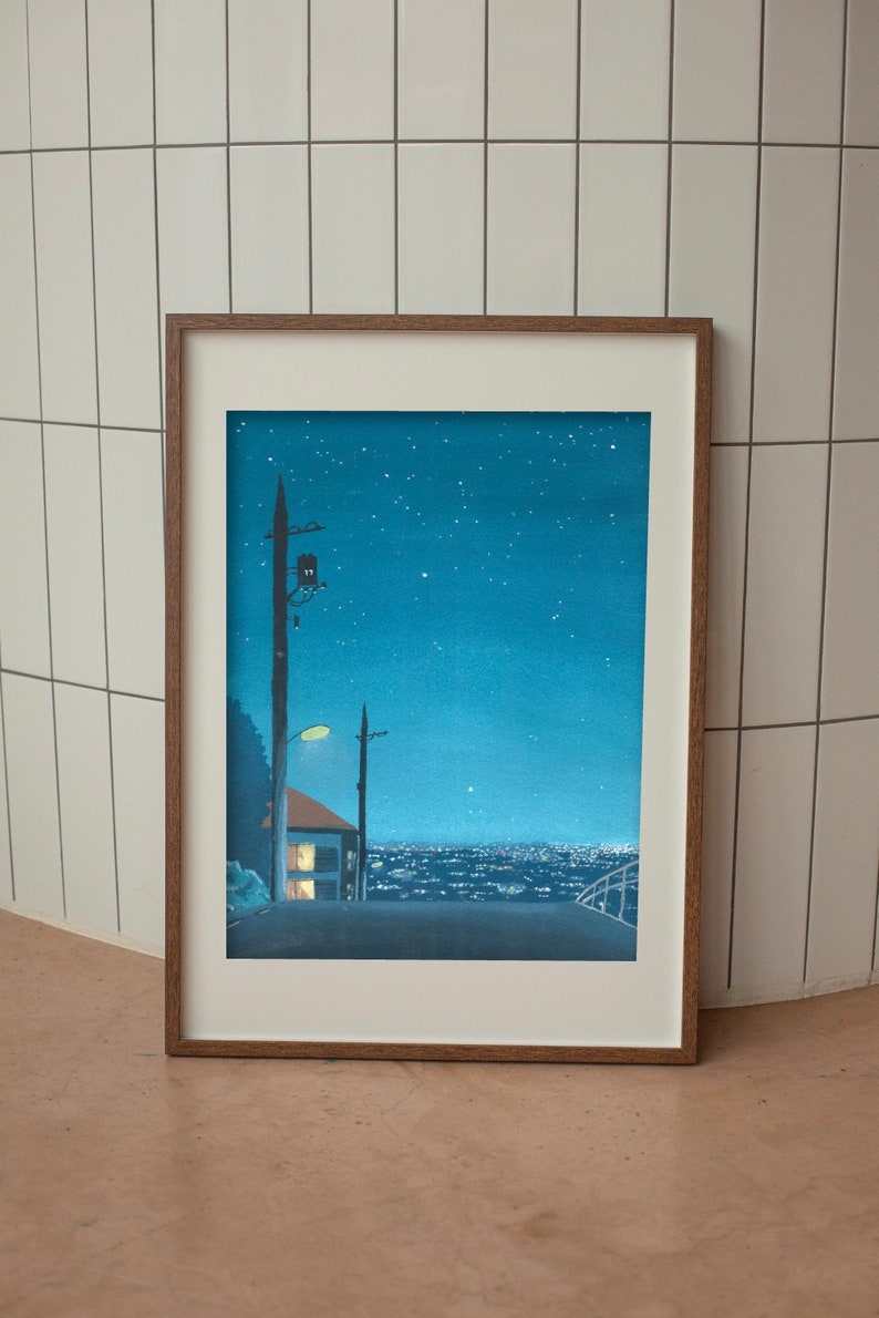 May include: Framed art print depicting a nighttime cityscape under a starry sky. The artwork features a road leading towards a town with illuminated buildings. A house with lit windows and utility poles are in the foreground. The frame is a warm brown color.