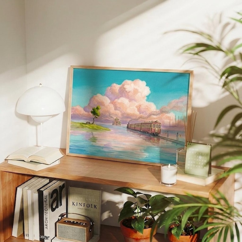 May include: A framed art print depicts a train traveling across water towards a distant city, under a sky filled with pink clouds. The artwork is displayed on a wooden shelf with books, a lamp, and a small potted plant. A candle and a calendar are also on the shelf.