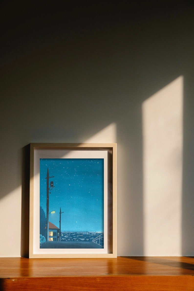 May include: Framed art print depicting a nighttime cityscape under a starry sky. The artwork features a street with a lamppost, a house with lit windows, and a distant city skyline. The print is in a light wooden frame.
