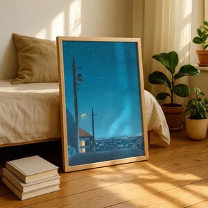 May include: A framed art print depicts a nighttime cityscape with a starry sky and a road leading into the distance. The artwork is in a light wooden frame and leans against a bed and a stack of books. The color palette is dominated by blues and greens.