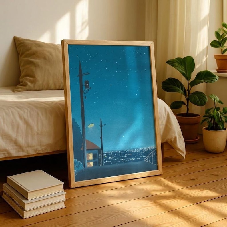 May include: A framed art print depicts a nighttime cityscape with a starry sky and a road leading into the distance. The artwork is in a light wooden frame and leans against a bed and a stack of books. The color palette is dominated by blues and greens.