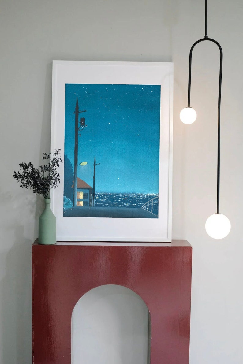 May include: A framed art print depicts a nighttime cityscape under a starry sky. A green vase with dark foliage sits on a red cabinet. A black, modern light fixture with two spherical bulbs hangs nearby.