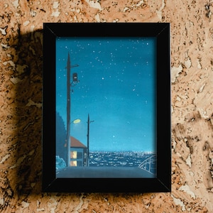May include: Framed art print depicting a nighttime cityscape under a starry sky. The artwork features a road, a house with lit windows, and utility poles. The frame is black, and the background is a textured, light brown surface.