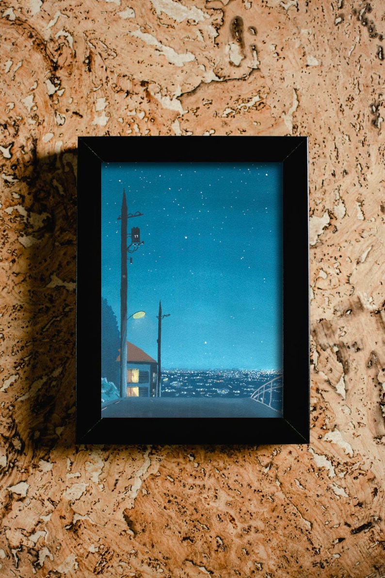 May include: Framed art print depicting a nighttime cityscape under a starry sky. The artwork features a road, a house with lit windows, and utility poles. The frame is black, and the background is a textured, light brown surface.