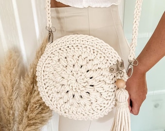 May include: A white crocheted round bag with a long white strap and a wooden bead tassel. The bag is worn over the shoulder of a person wearing white pants and a white top.