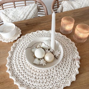 Puede incluir: Un mantel individual de crochet blanco con borde festoneado, que presenta un cuenco blanco lleno de adornos blancos y crema. El mantel individual est&aacute; sobre una mesa de madera con dos sillas de mimbre en el fondo.