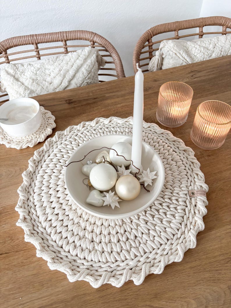 Puede incluir: Un mantel individual de crochet blanco con borde festoneado, que presenta un cuenco blanco lleno de adornos blancos y crema. El mantel individual est&aacute; sobre una mesa de madera con dos sillas de mimbre en el fondo.