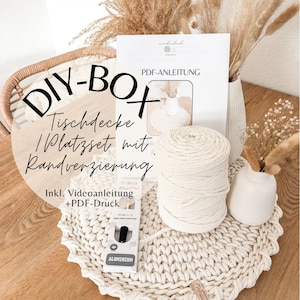 Könnte beinhalten: Eine DIY-Box mit dem Text "DIY-BOX Tischdecke / Platzset mit Randverzierung" enthält ein Häkelmuster, eine Kugel cremefarbener Wolle und einen Häkelhaken. Das Set enthält auch eine PDF-Anleitung und ein Video-Tutorial. Das fertige Produkt ist ein cremefarbener Platzset.