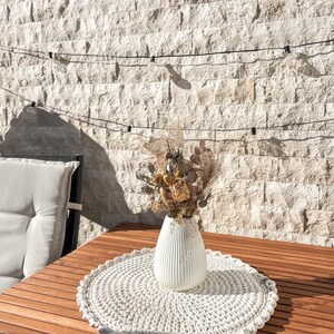 Puede incluir: Un camino de mesa de crochet blanco con un jarr&oacute;n de cer&aacute;mica blanco lleno de flores secas sobre una mesa de madera.