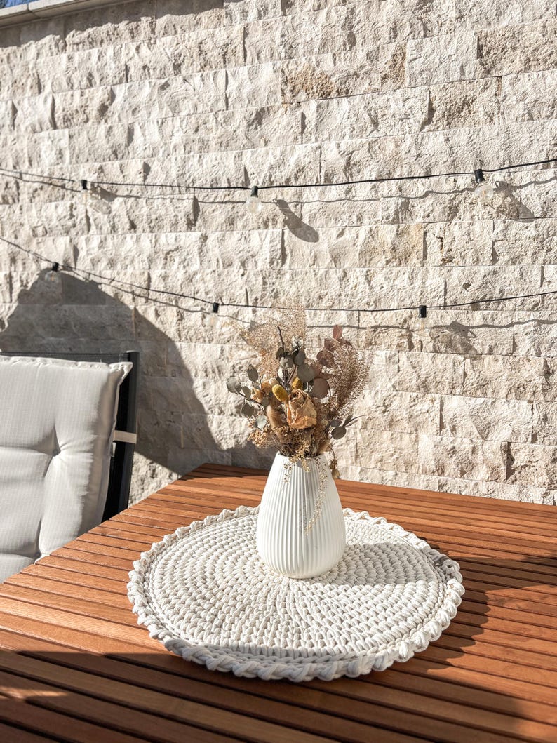 Puede incluir: Un camino de mesa de crochet blanco con un jarr&oacute;n de cer&aacute;mica blanco lleno de flores secas sobre una mesa de madera.