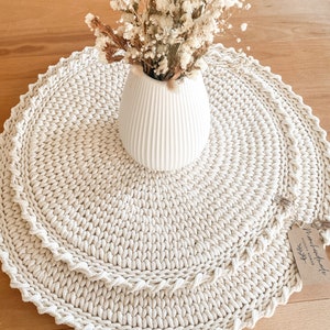 Könnte beinhalten: Eine beige runde Tischunterlage aus Häkelarbeit mit einer weißen Vase, gefüllt mit getrockneten weißen Blumen. Die Unterlage hat einen gewellten Rand und ist aus dickem Garn gefertigt. Ein kleines Schild mit dem Text "Handmade with love" ist an der Unterlage befestigt.