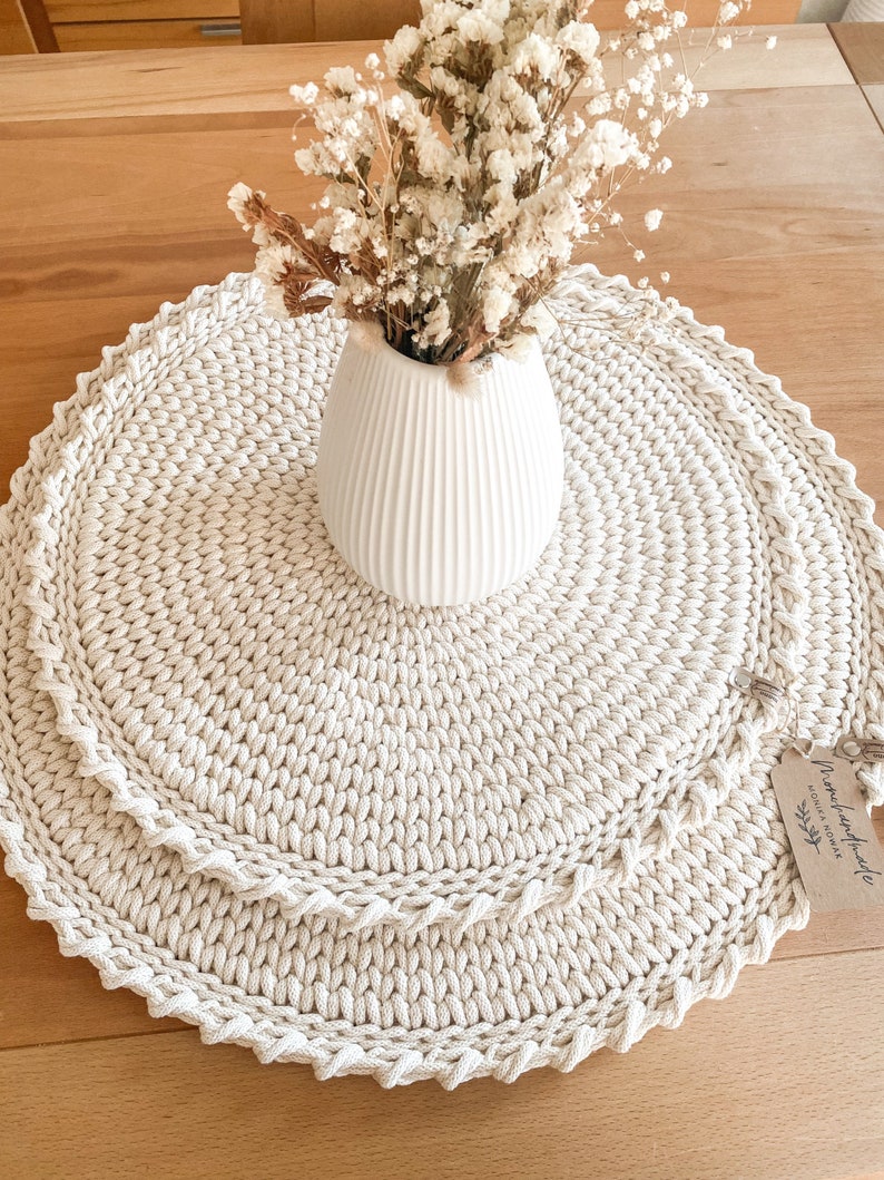 Puede incluir: Un mantel redondo de crochet beige con un jarr&oacute;n blanco lleno de flores blancas secas. El mantel tiene un borde festoneado y est&aacute; hecho de hilo grueso. Una peque&ntilde;a etiqueta con el texto "hecho a mano con amor" est&aacute; unida al mantel.