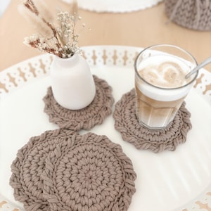Puede incluir: Una bandeja blanca con un pequeño jarrón blanco con flores secas, un vaso de café con leche y varios posavasos de ganchillo marrones. Los posavasos son circulares con un borde trenzado texturizado.