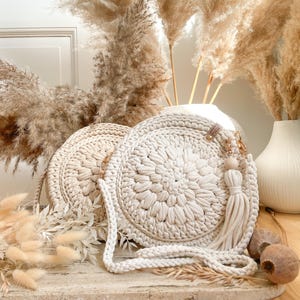 K&ouml;nnte beinhalten: Zwei cremefarbene runde geh&auml;kelte Taschen mit einem langen cremefarbenen Riemen. Die Taschen haben ein Quasten-Detail und liegen auf einer hellbraunen Holzoberfl&auml;che mit getrocknetem Pampasgras im Hintergrund.