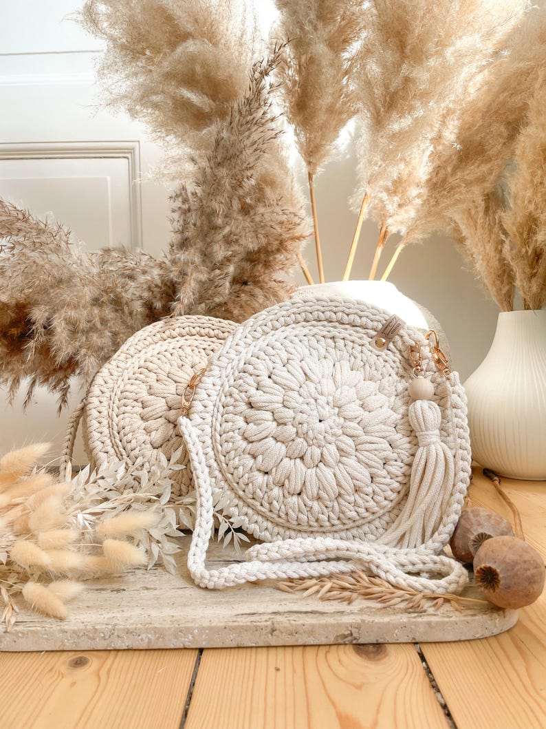 K&ouml;nnte beinhalten: Zwei cremefarbene runde geh&auml;kelte Taschen mit einem langen cremefarbenen Riemen. Die Taschen haben ein Quasten-Detail und liegen auf einer hellbraunen Holzoberfl&auml;che mit getrocknetem Pampasgras im Hintergrund.