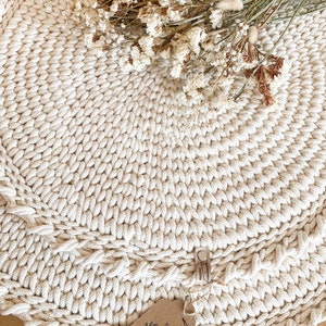 Puede incluir: Una alfombra redonda de crochet de color crema con una textura de fibra natural. La alfombra est&aacute; hecha con cuerda gruesa y tiene un patr&oacute;n decorativo. Un ramo de flores secas se coloca encima de la alfombra. La alfombra tiene una etiqueta que dice "Hecho a mano con amor" y "MONIK & NOMAX".