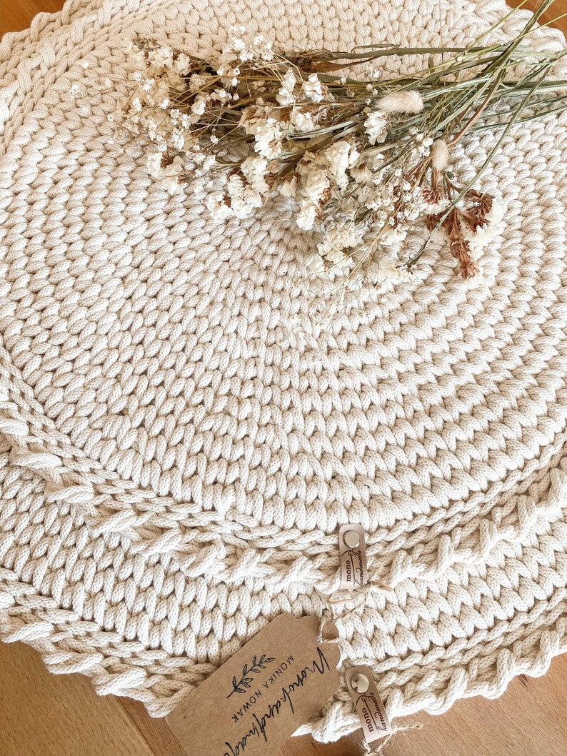 Puede incluir: Una alfombra redonda de crochet de color crema con una textura de fibra natural. La alfombra est&aacute; hecha con cuerda gruesa y tiene un patr&oacute;n decorativo. Un ramo de flores secas se coloca encima de la alfombra. La alfombra tiene una etiqueta que dice "Hecho a mano con amor" y "MONIK & NOMAX".