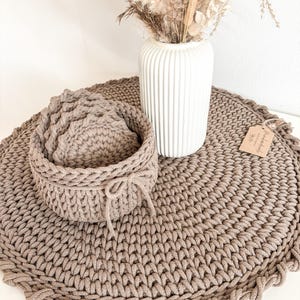 Puede incluir: Un juego de cuatro posavasos de crochet marr&oacute;n con un cuenco de crochet marr&oacute;n a juego. Los posavasos y el cuenco est&aacute;n sobre una alfombra redonda de crochet marr&oacute;n grande. Un jarr&oacute;n blanco con flores secas est&aacute; en el fondo.