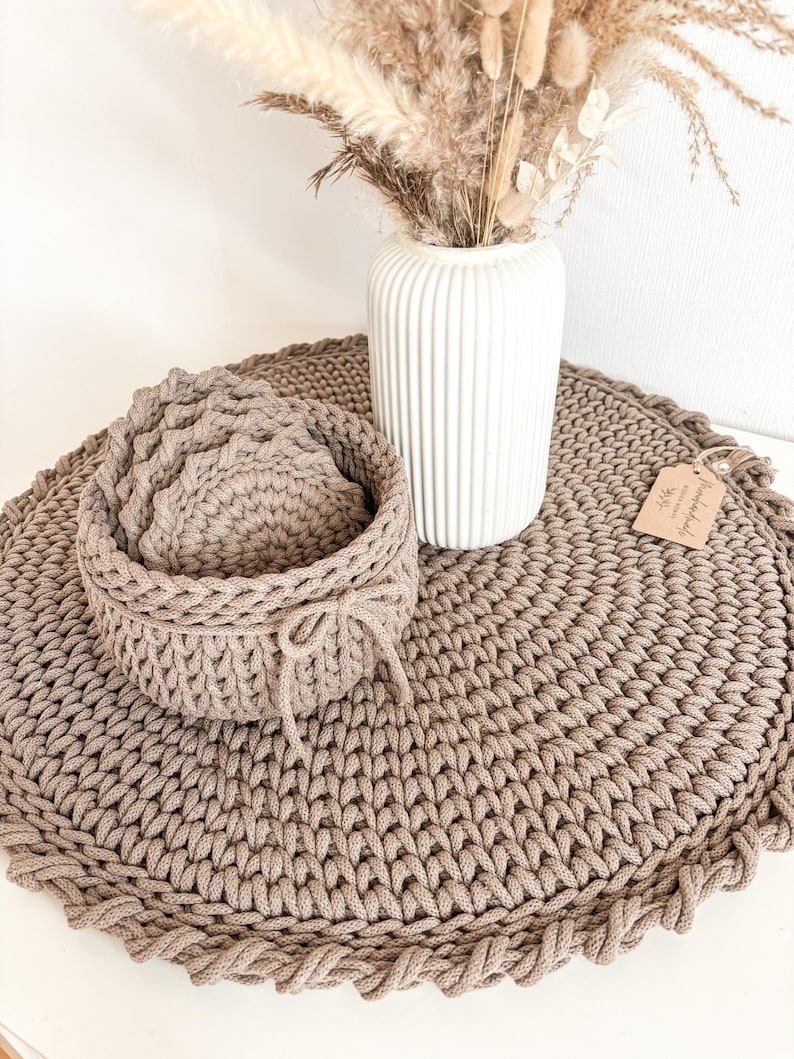 Puede incluir: Un juego de cuatro posavasos de crochet marr&oacute;n con un cuenco de crochet marr&oacute;n a juego. Los posavasos y el cuenco est&aacute;n sobre una alfombra redonda de crochet marr&oacute;n grande. Un jarr&oacute;n blanco con flores secas est&aacute; en el fondo.