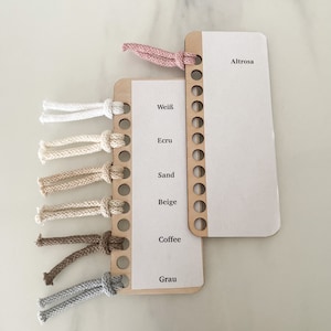 Puede incluir: Un muestrario de hilo de madera con diferentes colores de hilo enrollados alrededor. Los colores son: blanco, crudo, arena, beige, caf&eacute; y gris. El muestrario tiene las palabras "Wei&szlig;", "Ecru", "Sand", "Beige", "Coffee" y "Grau" impresas en &eacute;l. Hay un segundo muestrario con la palabra "Altrose" impresa en &eacute;l.