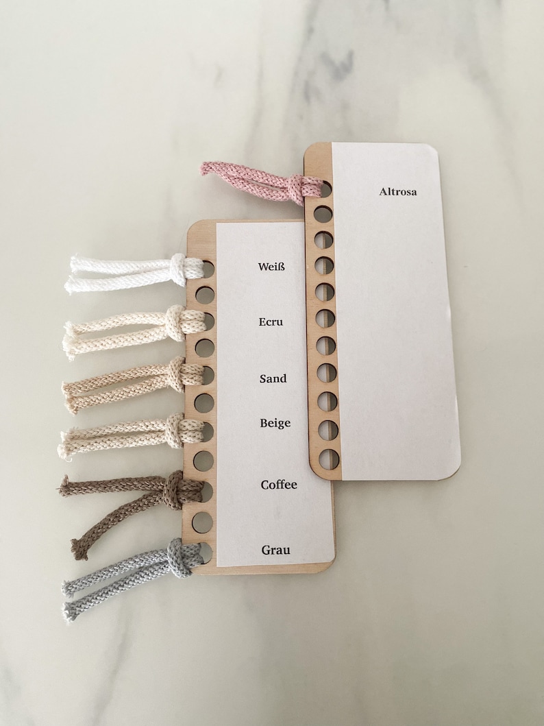 Puede incluir: Un muestrario de hilo de madera con diferentes colores de hilo enrollados alrededor. Los colores son: blanco, crudo, arena, beige, caf&eacute; y gris. El muestrario tiene las palabras "Wei&szlig;", "Ecru", "Sand", "Beige", "Coffee" y "Grau" impresas en &eacute;l. Hay un segundo muestrario con la palabra "Altrose" impresa en &eacute;l.