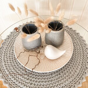 Puede incluir: Dos jarrones de cer&aacute;mica gris con hierba de la pampa seca sobre un camino de mesa redondo de crochet gris. Una figurita de p&aacute;jaro de cer&aacute;mica blanca est&aacute; sobre un plato de cer&aacute;mica beige.
