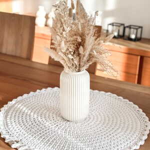 Puede incluir: Un camino de mesa redondo de crochet blanco con un jarr&oacute;n de cer&aacute;mica blanco con hierba de la pampa seca y otras flores secas.