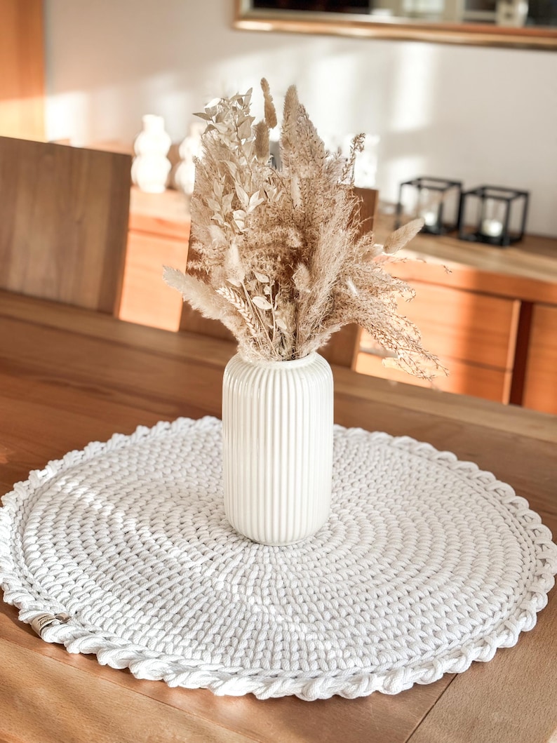 Puede incluir: Un camino de mesa redondo de crochet blanco con un jarr&oacute;n de cer&aacute;mica blanco con hierba de la pampa seca y otras flores secas.