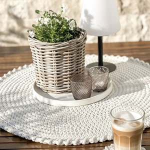 Puede incluir: Un camino de mesa redondo de crochet blanco con una bandeja de cer&aacute;mica blanca, una maceta de mimbre con flores blancas, dos candelabros de vidrio transparente y un vaso de caf&eacute; sobre un posavasos de crochet blanco sobre una mesa de madera.