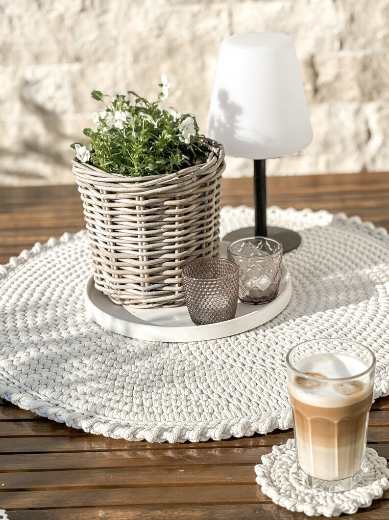 Puede incluir: Un camino de mesa redondo de crochet blanco con una bandeja de cer&aacute;mica blanca, una maceta de mimbre con flores blancas, dos candelabros de vidrio transparente y un vaso de caf&eacute; sobre un posavasos de crochet blanco sobre una mesa de madera.