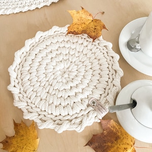 Könnte beinhalten: Handgefertigter, cremefarbener Häkel-Platzdeckchen mit gewelltem Rand. Der runde Platzdeckchen liegt auf einer hellen Holzoberfläche, mit einem Lederanhänger. Herbstblätter und eine Tasse mit Untertasse sind ebenfalls vorhanden.